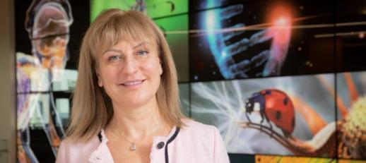 Blonde woman with pink jacket standing in front of a collage wall of science themed pictures